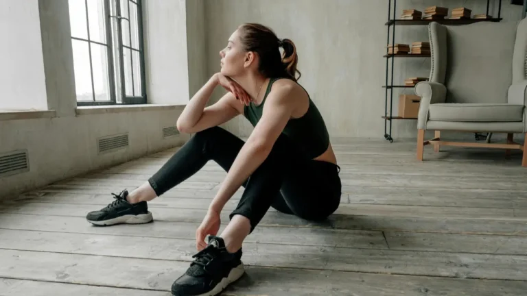 Kobieta ubrana w sportowe odzienie siedzi na podłodze w domu i zastanawia się, jaki sprzęt kupić do domowej siłowni
